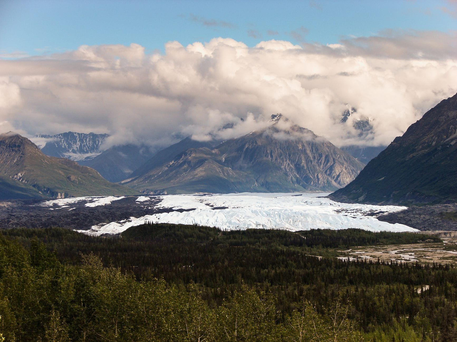 Wide view of Matanuska Glacier flowing through a forested valley beneath cloud-covered Alaskan mountains.